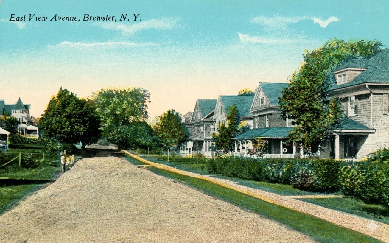 Early-twentieth-century color picture postcard captioned 'East View Avenue, Brewster, N.Y.,' showing a dirt road lined with houses; a turreted Victorian sits at the left.