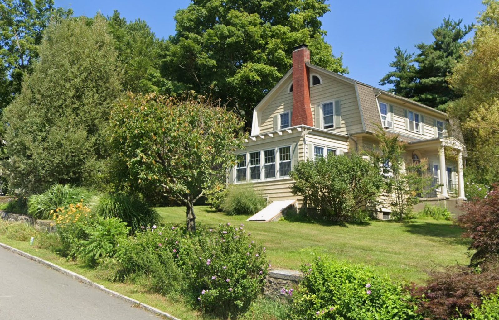 Modern color photograph of a cream-colored gambrel-roofed Dutch Colonial house with green shutters, a red-brick central chimney, a half-round attic window, and an enclosed sun porch, set behind a lawn and garden.