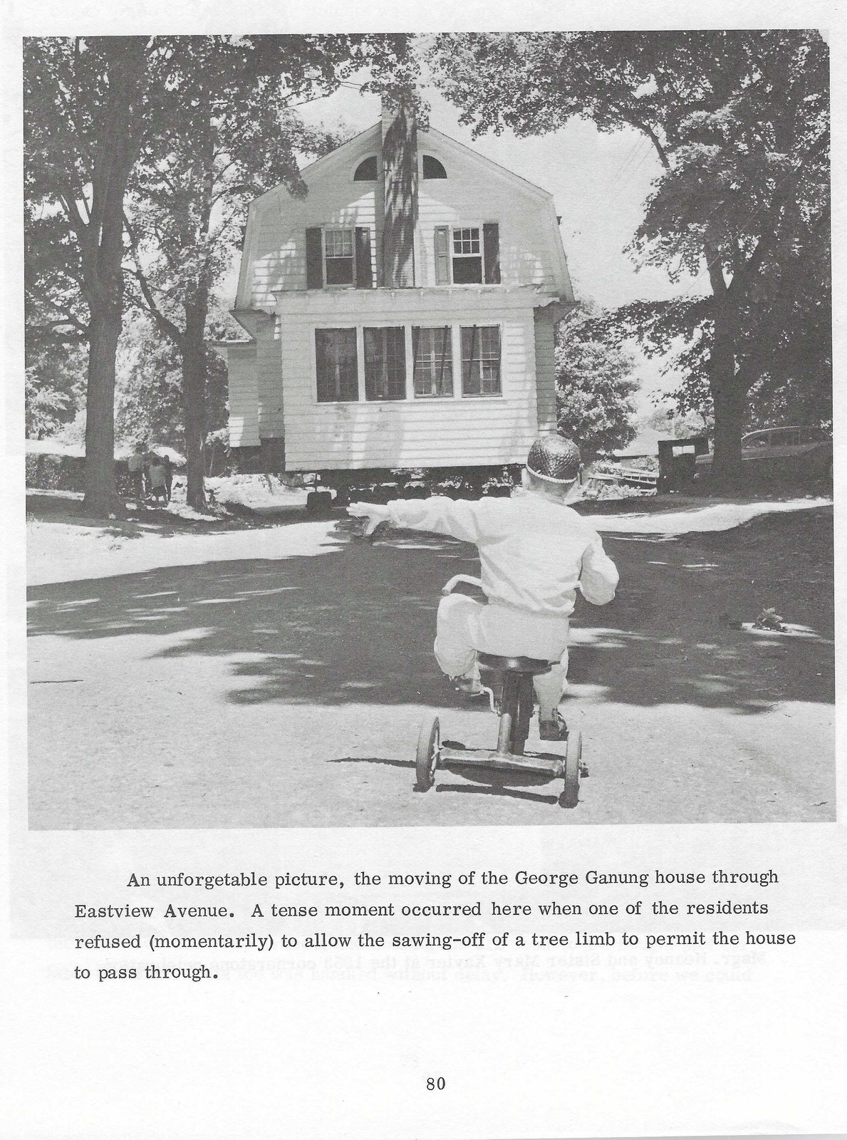 Black-and-white photograph of a gambrel-roofed Dutch Colonial house with a half-round attic window and central chimney, raised on cribbing and being moved along a street, with a small child on a tricycle watching from the foreground.