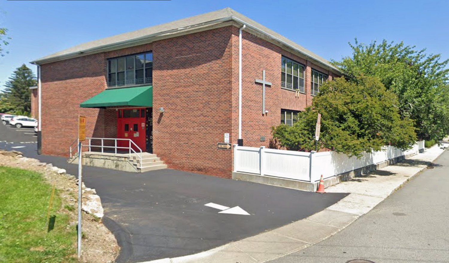 Color photograph of the corner of a two-story brick school building with a cross mounted on the wall, a green awning over a red door and a railed entrance ramp, at a street corner on Eastview Avenue.