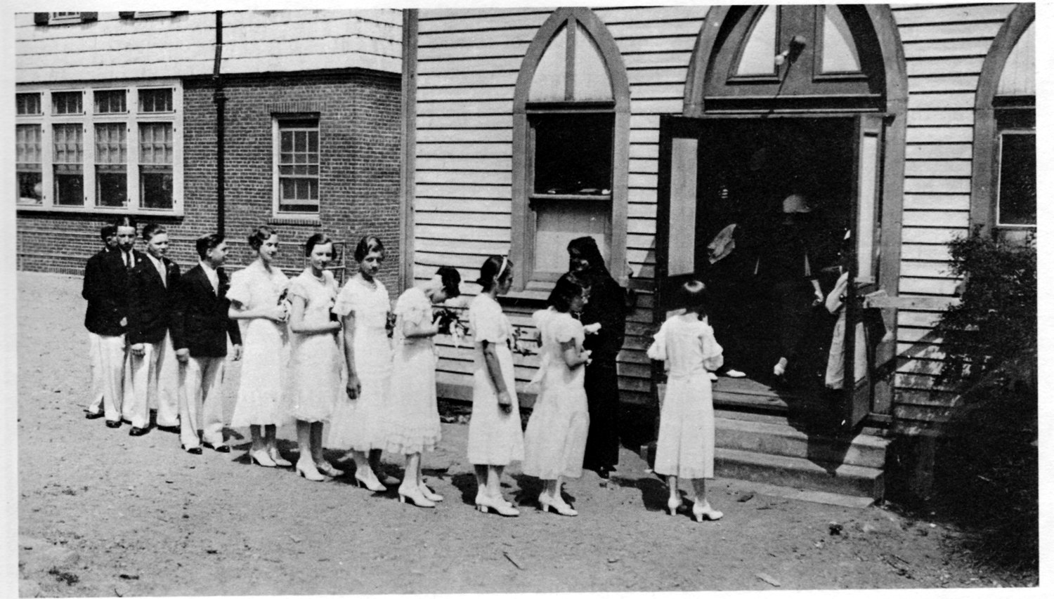 1930s photograph of school graduates in white dresses and dark suits filing into the arched doorway of the old wooden church, a nun and priest at the door, the brick parochial school visible behind at left.