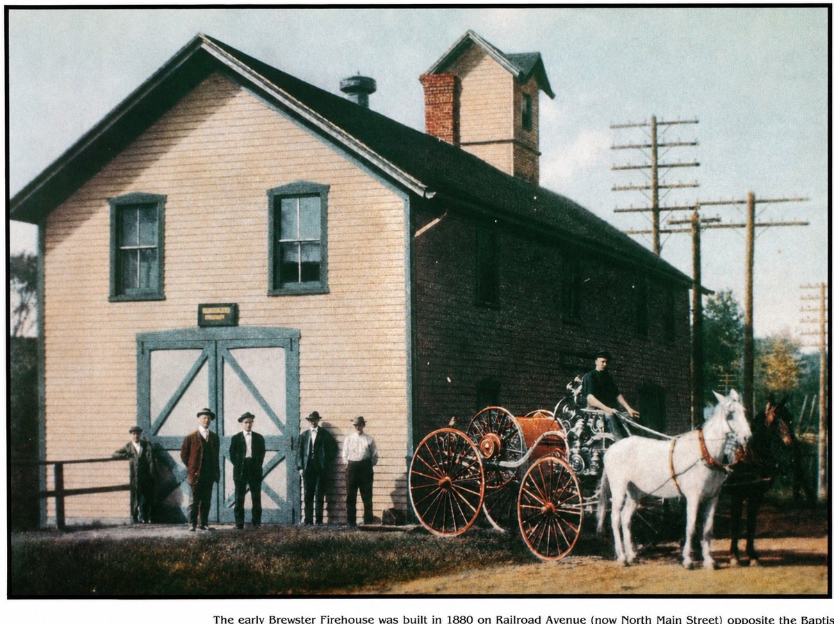 The original 1880 Brewster Firehouse: a two-story yellow clapboard building with a steeply pitched roof and a tall hose-drying tower with a brick chimney. The front of the building has wide blue double doors with diagonal bracing under a small signboard. Five men in dark suits and hats stand by the doors. To the right, a single firefighter sits on a horse-drawn fire engine pulled by two white horses, with the apparatus visible in red and brass. Telegraph poles and trees in the background; this is an early-20th-century photograph, AI-colorized.