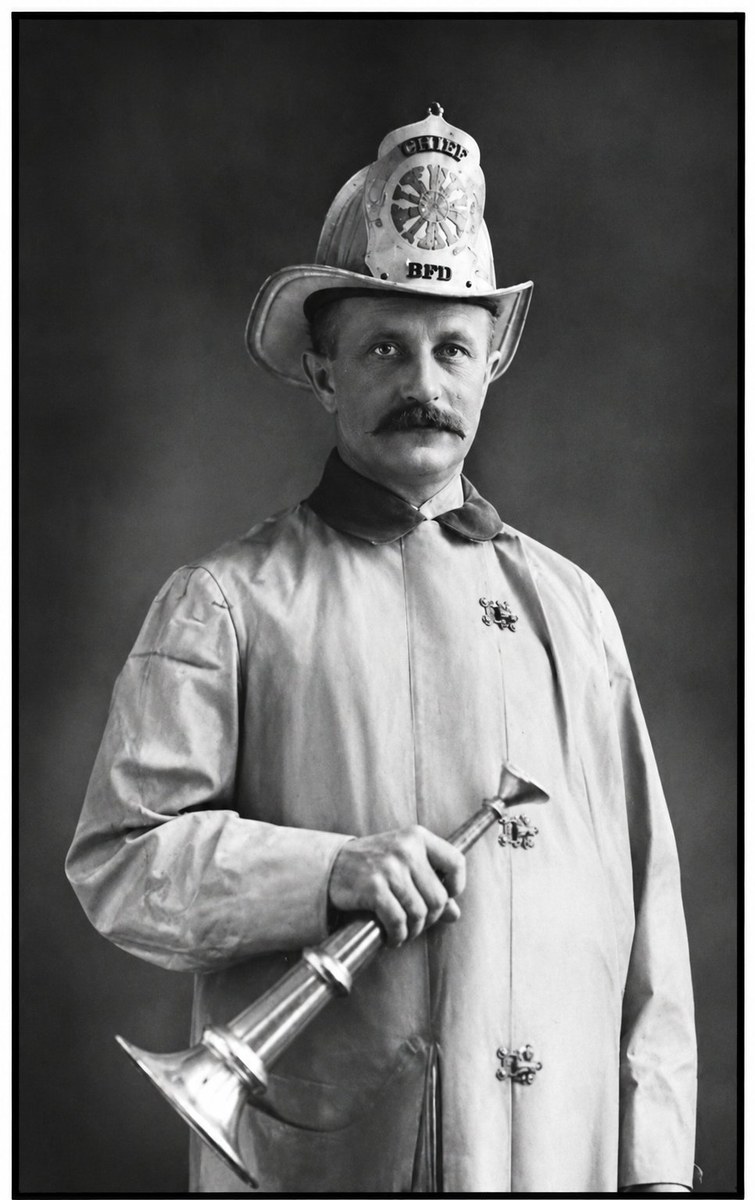 Formal studio portrait of John D. Morehouse in fire-chief regalia: white leather chief's helmet lettered 'CHIEF' above and 'BFD' below a wheel-shaped front shield; light-colored full-length canvas fire coat with metal clasps; holding a silver speaking trumpet across his chest. He has a brushed-up mustache and looks directly at the camera, mid-thirties to forties.