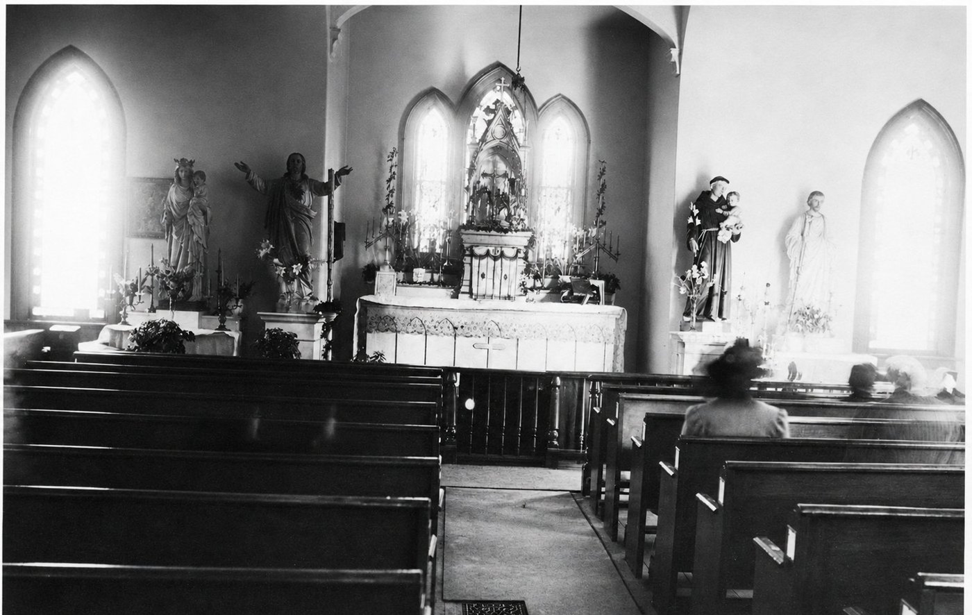 Interior photograph of a small Gothic-windowed wooden church in the 1890s: a central altar with statuary and flowers beneath three lancet windows, side altars of the Virgin and St. Joseph, dark wooden pews, a few women seated at right.