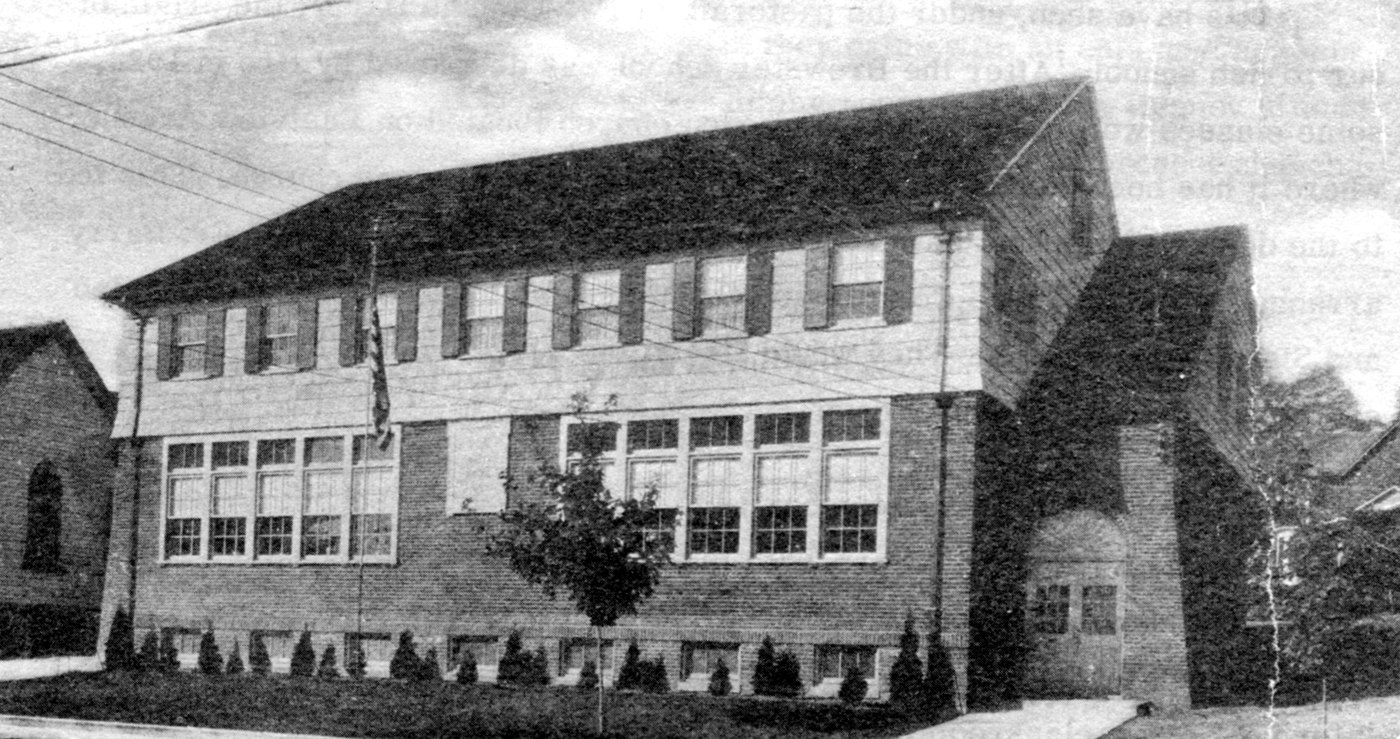 Black-and-white photograph of a two-story brick parochial school with a hipped roof, large multi-pane classroom windows and a corner entrance bay, young shrubs along the foundation; the gable of a brick church visible at the left edge.