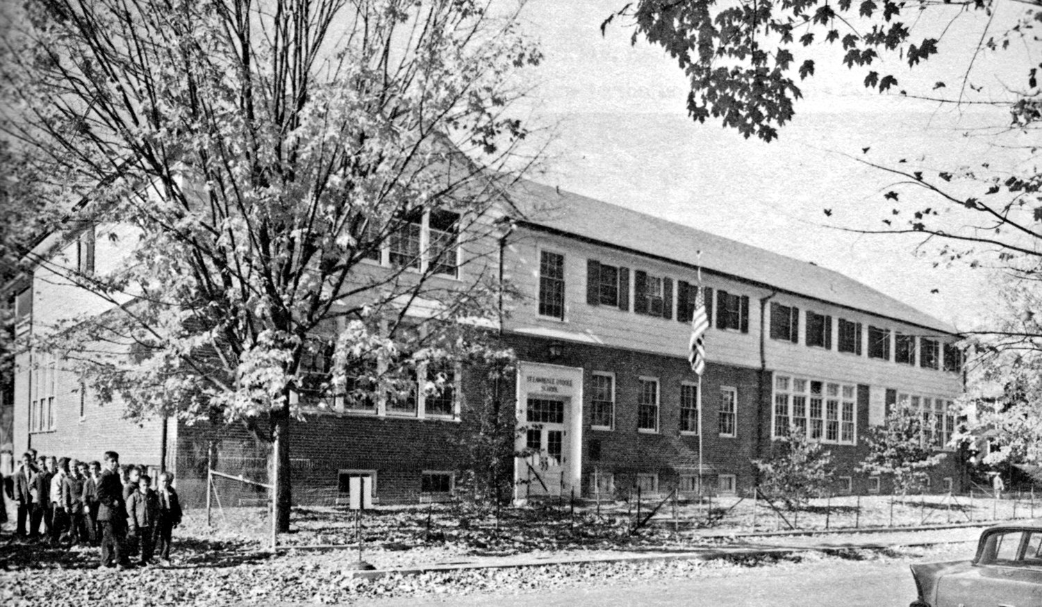 Black-and-white photograph of the enlarged brick school of 1953, a long two-story range with a corner entrance lettered 'St. Lawrence O'Toole School,' a 1950s automobile at right and a group of schoolchildren at left under autumn trees.
