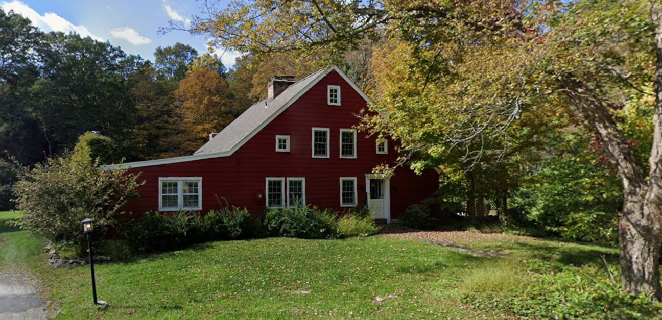 The Gage House today: a red-painted clapboard saltbox farmhouse with a steep wood-shingled roof, white-trimmed windows, and an open front lawn shaded by mature trees in fall foliage.