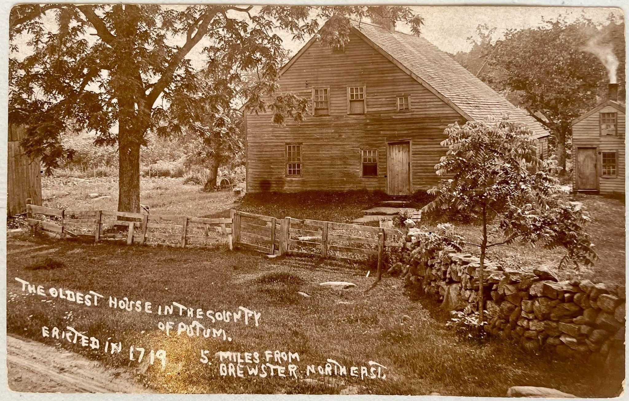 Sepia-toned historic postcard showing the Gage House, a saltbox-form farmhouse with steep roof, surrounded by trees, rail fences, and a stone wall. Inscription at bottom: THE OLDEST HOUSE IN THE COUNTY OF PUTNAM. ERECTED IN 1719. 5 MILES FROM BREWSTER NORTHEAST.