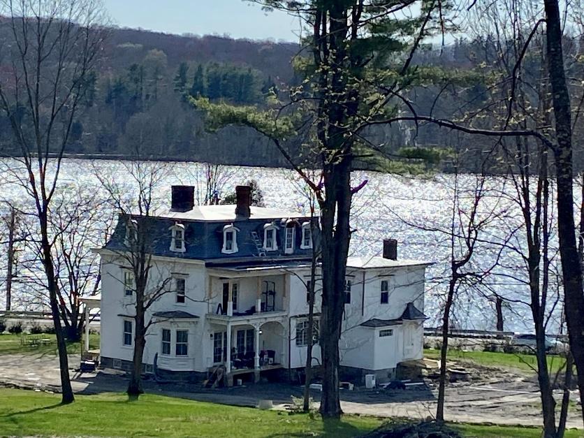 The house under restoration in spring 2024, seen from across the Middle Branch Reservoir — white clapboard French Second Empire structure with mansard roof, three chimneys, surrounded by bare-branched trees.