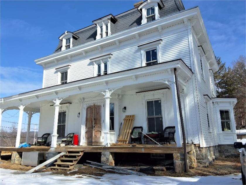 Close-up of the house porch and front facade under restoration, showing fresh white paint, removed door, scaffolding and lumber on the porch deck.