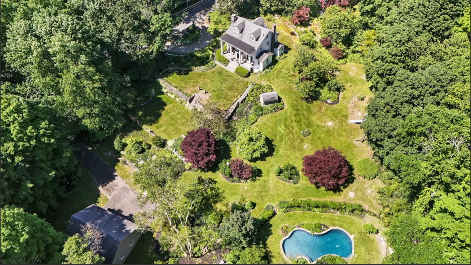 Aerial view of the Hammarskj&ouml;ld House property: the white farmhouse sits at top center, fronted by a columned porch and reached by a driveway from the lower left. Below and around it spread terraced lawns held by stone retaining walls, ornamental Japanese maples, perennial beds, a small outbuilding, and a kidney-shaped swimming pool at lower right, all enclosed by mature woodland.
