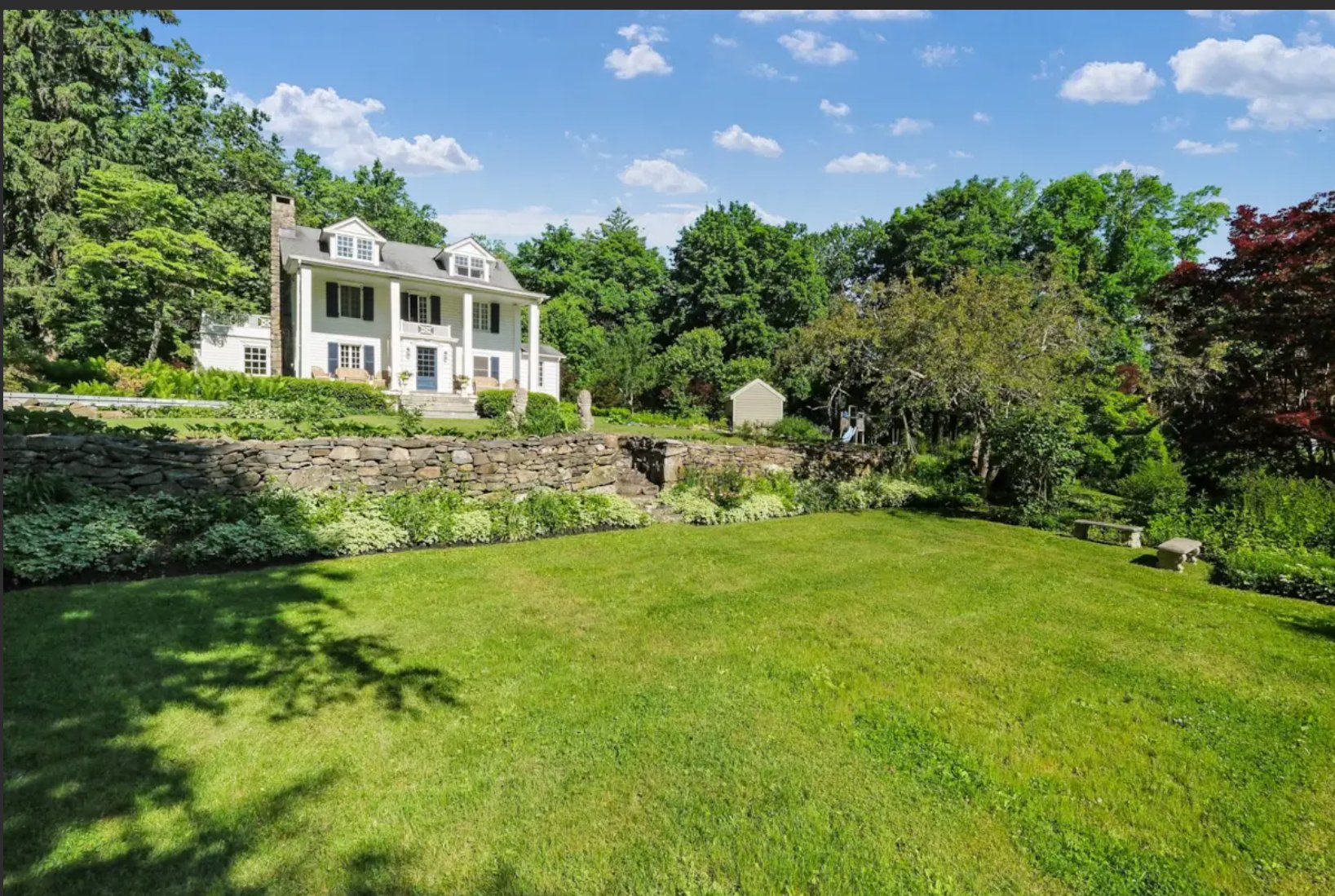 The Hammarskj&ouml;ld House seen from the lawn below, with a long dry-laid fieldstone retaining wall running across the foreground and the white Greek Revival farmhouse rising on the terrace above it. A small shed stands to the right; stone garden benches sit at the edge of the lawn.