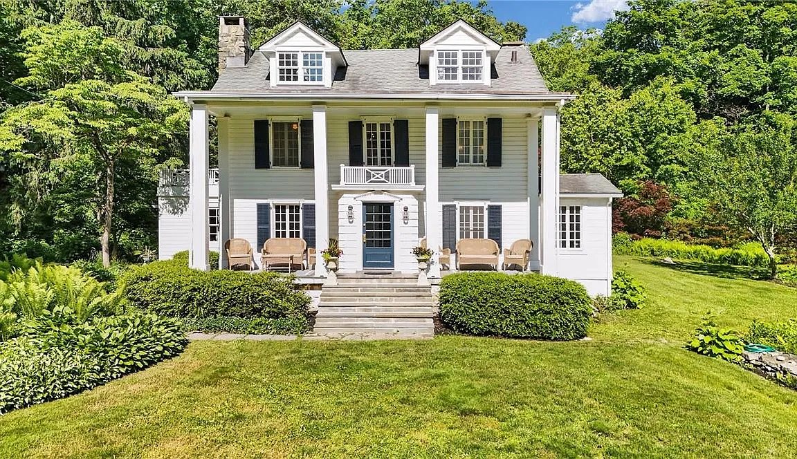 A two-story white-clapboard Greek Revival farmhouse with a columned center-entry portico, two roof dormers with multi-pane windows, blue shutters, and a small second-story balcony over the front door. The house sits behind a low stone wall and a foundation planting of shrubs, with mature trees framing it on either side.