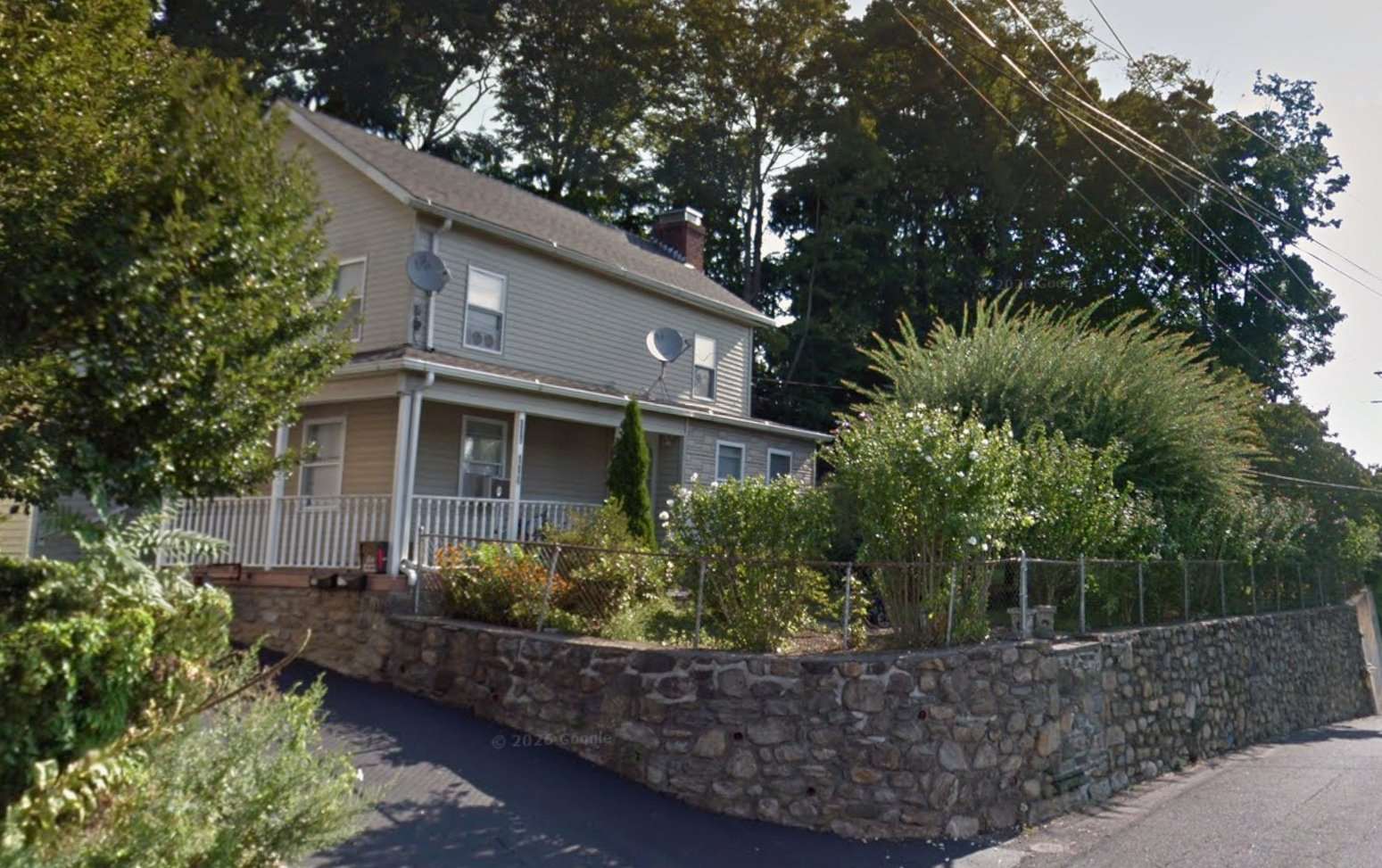 Google Street View photograph of the house at 110-114 Oak Street: a cream-painted vernacular wood-frame house with side-gabled roof and full-width front porch, set above a long hand-laid fieldstone retaining wall that wraps the corner along the road.