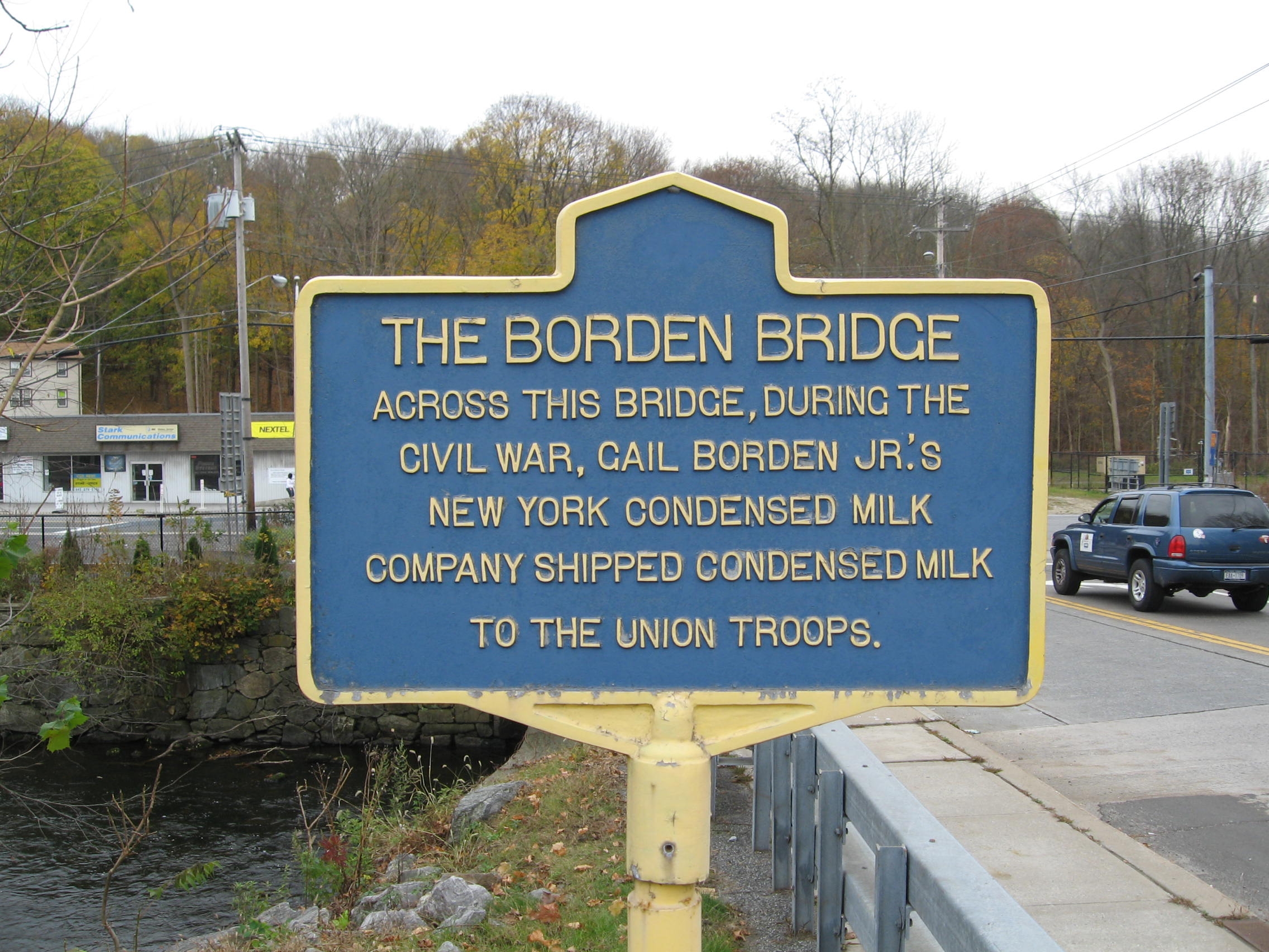 Blue and yellow New York State-style historic marker reading THE BORDEN BRIDGE. Across this bridge, during the Civil War, Gail Borden Jr.'s New York Condensed Milk Company shipped condensed milk to the Union Troops. The marker stands at the edge of a small bridge on Main Street in the Village of Brewster, with the East Branch of the Croton River visible to its left and Main Street traffic passing to its right.