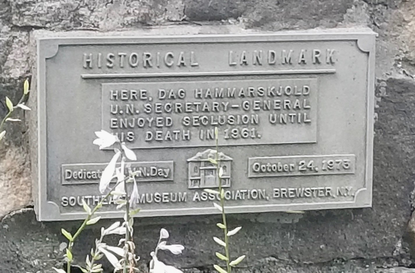Cast metal plaque, in close-up, reading HISTORICAL LANDMARK at the top, then in a recessed central panel: Here, Dag Hammarskjold U.N. Secretary-General enjoyed seclusion until his death in 1961. Below this, at lower left, a small box reading Dedicated on U.N. Day; at center a small relief image of a two-story house with a columned center entry and dormer windows; and at lower right October 24, 1973. Across the bottom: SOUTHEAST MUSEUM ASSOCIATION, BREWSTER, N.Y.