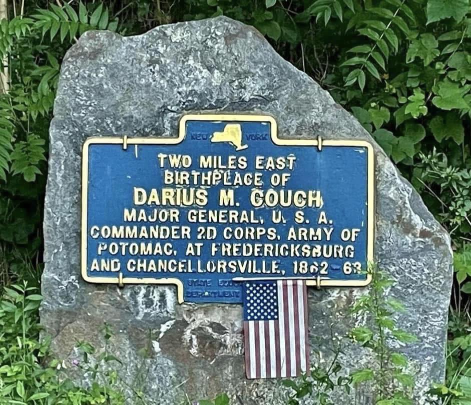 Blue and yellow New York State historic marker mounted on a large boulder reading TWO MILES EAST: Birthplace of Darius M. Couch, Major General U.S.A., Commander 2D Corps Army of Potomac, at Fredericksburg and Chancellorsville, 1862-63. State Education Department. A small American flag has been planted at the base of the marker.