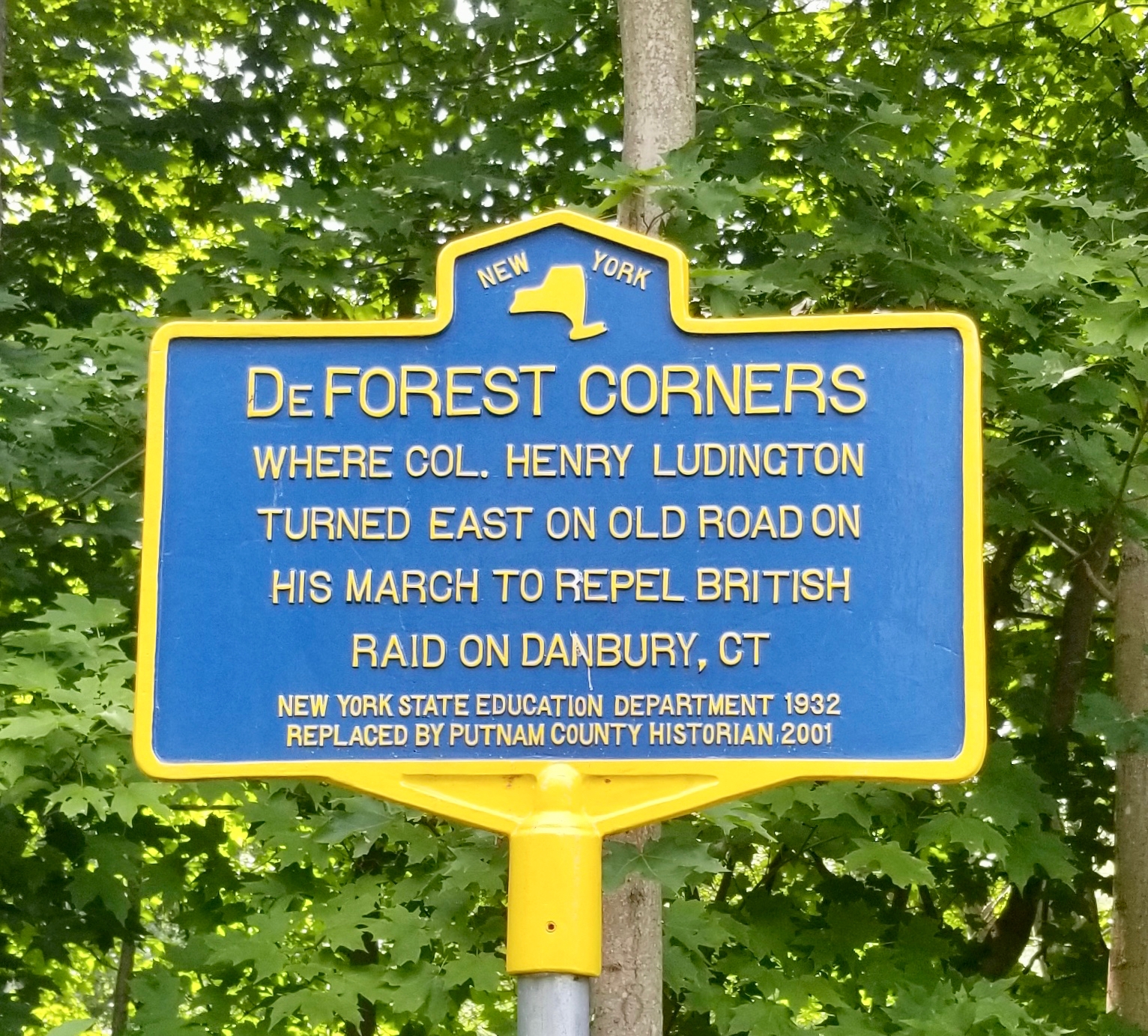 Blue and yellow New York State historic marker reading DeFOREST CORNERS. Where Col. Henry Ludington turned east on old road on his march to repel British raid on Danbury, CT. New York State Education Department 1932. Replaced by Putnam County Historian 2001.