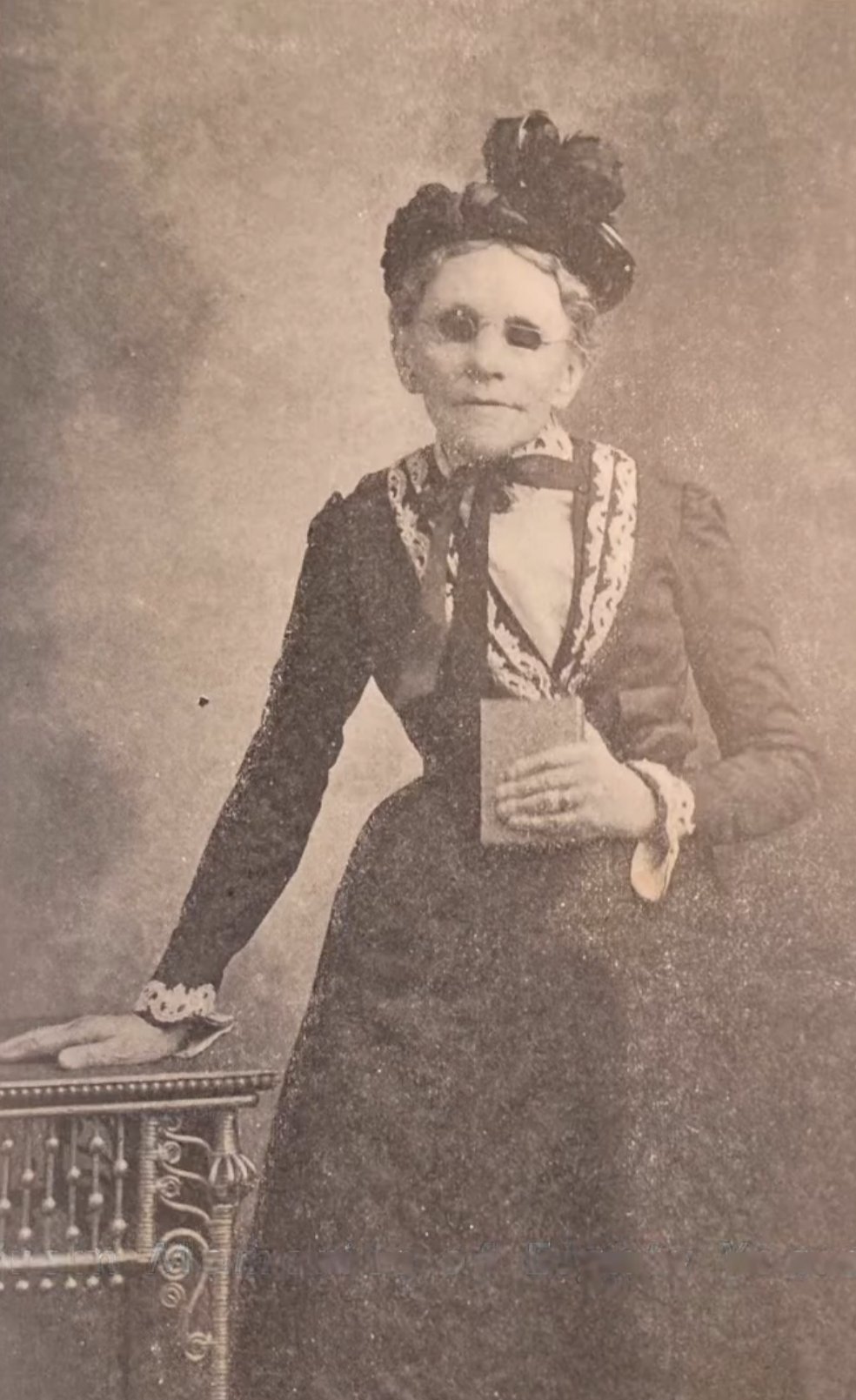 Sepia-toned photograph of Fanny Crosby in old age, standing beside a wicker table, wearing a dark dress with elaborate lace trim, an ornate feathered hat, and her characteristic dark glasses. She holds a small book — most likely a hymnal or Bible — in her left hand.