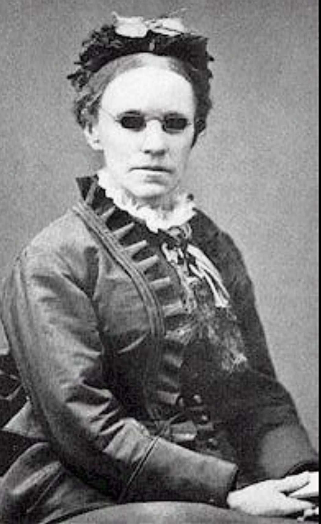 Black-and-white photograph of Fanny Crosby in middle age, seated, wearing a dark Victorian dress with a high white lace collar, ribbon trim, and a small ornate hat. Her dark glasses are visible; her expression is composed.