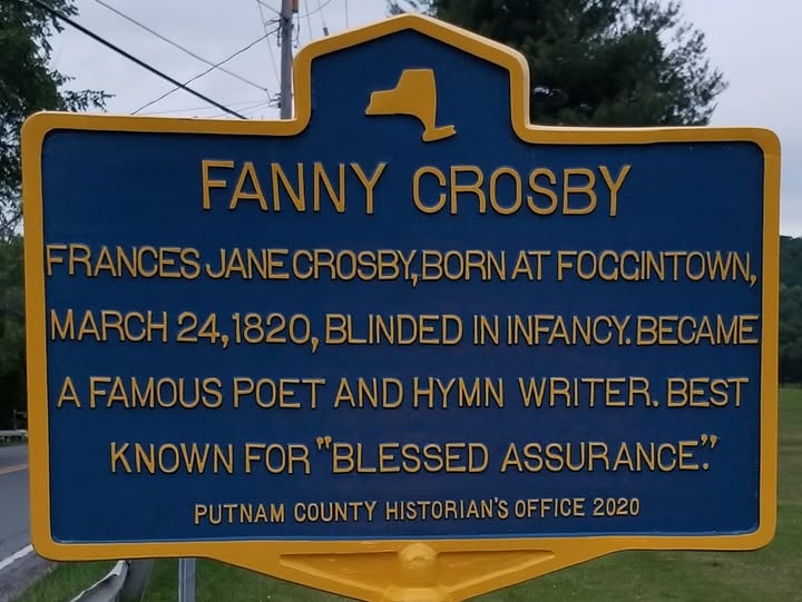 Blue and yellow New York State-style historic marker reading FANNY CROSBY: Frances Jane Crosby, born at Foggintown, March 24, 1820, blinded in infancy. Became a famous poet and hymn writer. Best known for 'Blessed Assurance.' Erected by the Putnam County Historian's Office in 2020.