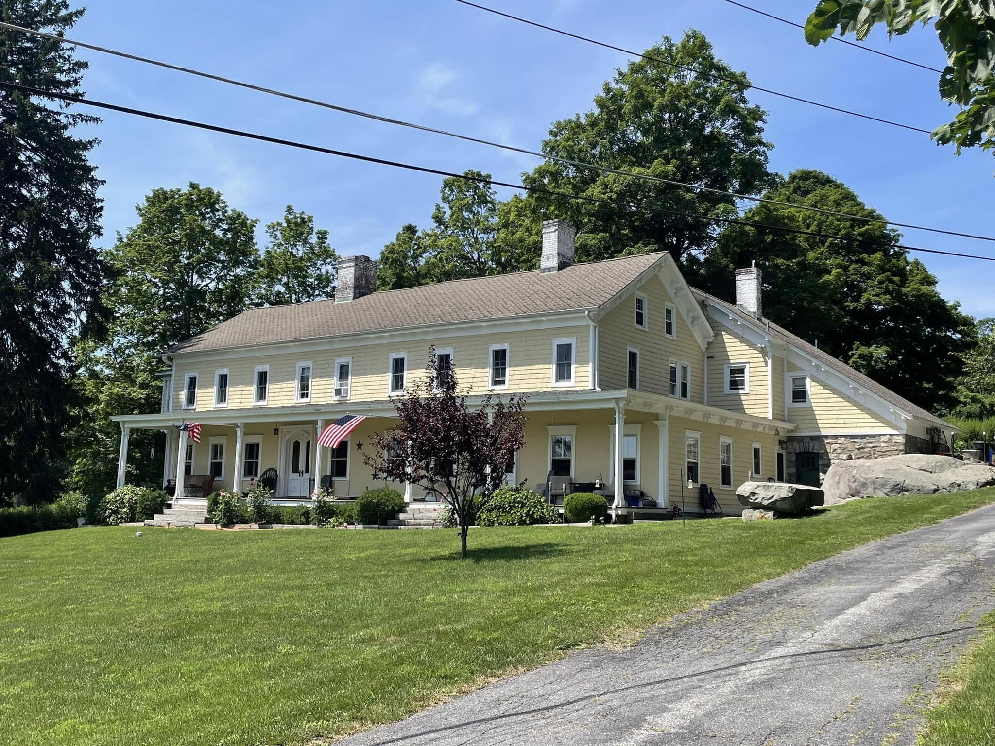 The Fowler House in summer: a long pale-yellow clapboard farmhouse with a full-width covered front porch, two American flags hanging from the porch posts, two brick chimneys rising from a steeply pitched roof, and a later two-story side wing extending to the right. A large granite outcrop is visible at the far right. The house sits behind a deep lawn at the end of a paved driveway, framed by mature shade trees.