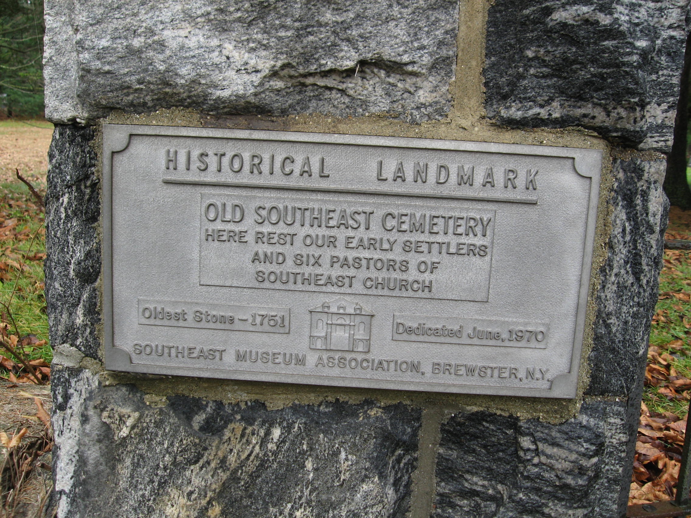 Cast metal plaque mounted on a rough stone pillar at the cemetery entrance, reading HISTORICAL LANDMARK at top. Below: OLD SOUTHEAST CEMETERY — HERE REST OUR EARLY SETTLERS AND SIX PASTORS OF SOUTHEAST CHURCH. At lower left: Oldest Stone — 1751. At center: a small relief image of a small church building with arched windows. At lower right: Dedicated June, 1970. Across the bottom: SOUTHEAST MUSEUM ASSOCIATION, BREWSTER, N.Y.