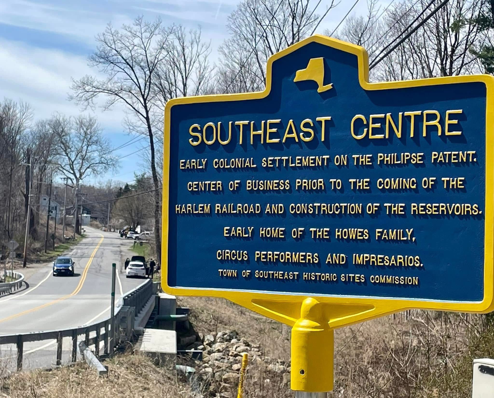 Blue and yellow New York State-style historic marker reading SOUTHEAST CENTRE, with inscription describing it as an early colonial settlement on the Philipse Patent, center of business prior to the Harlem Railroad and construction of the reservoirs, and early home of the Howes family, circus performers and impresarios.