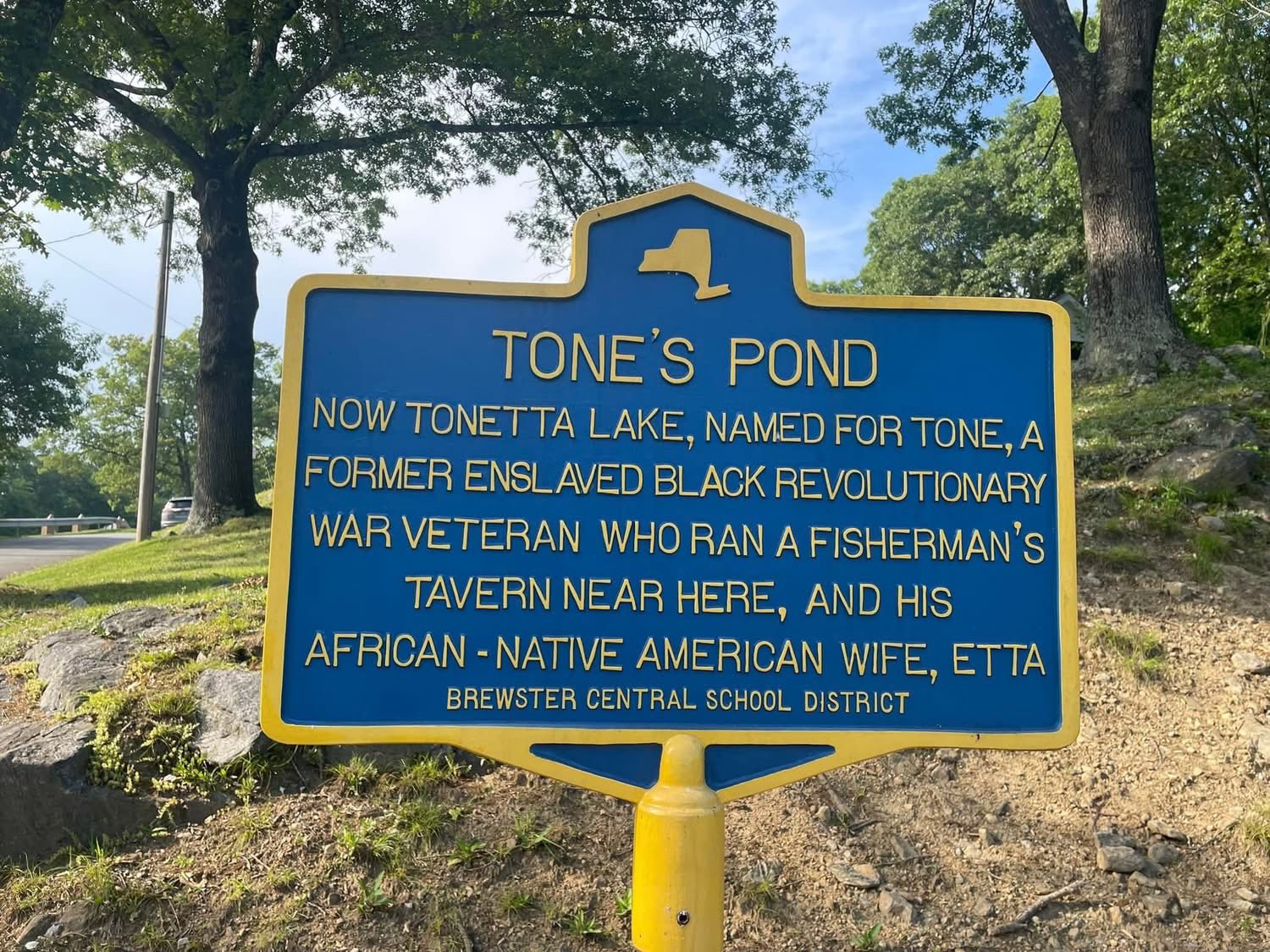 Blue and yellow New York State-style historic marker reading TONE'S POND. Now Tonetta Lake, named for Tone, a former enslaved Black Revolutionary War veteran who ran a fisherman's tavern near here, and his African-Native American wife, Etta. Brewster Central School District. The marker stands on a rise above the lake with autumn foliage in the background.