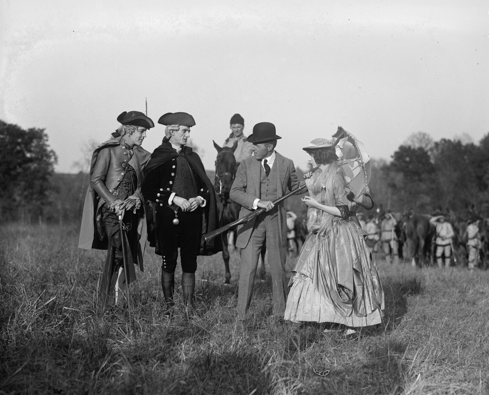 Black-and-white photograph in an open field: two men in tricorne hats, wigs, and Revolutionary-era coats and capes stand at left; a mounted rider waits behind them; a man in a business suit and bowler hat holds a flintlock musket at center; a young woman in an eighteenth-century gown with a parasol stands at right; a line of costumed extras and horses is drawn up in the distance.