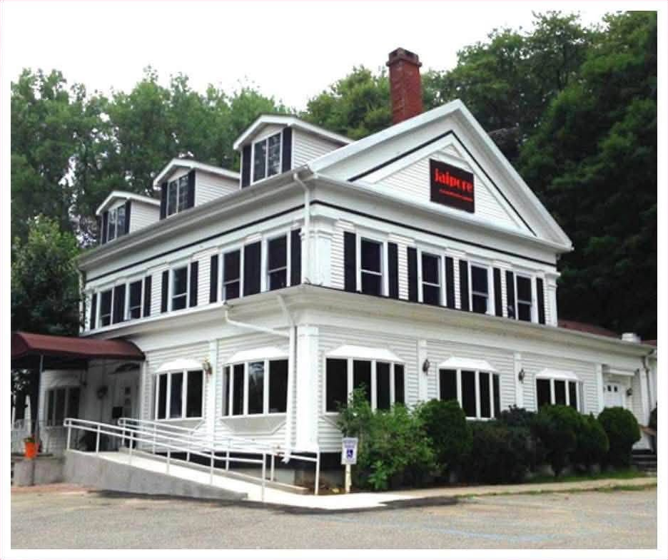 Large white three-story gable-front house with black shutters, boxed cornice, corner pilasters, awninged porch windows and a tall brick chimney, standing among trees — the former Theall homestead, now the Jaipore Royal Indian Cuisine restaurant on Route 22.