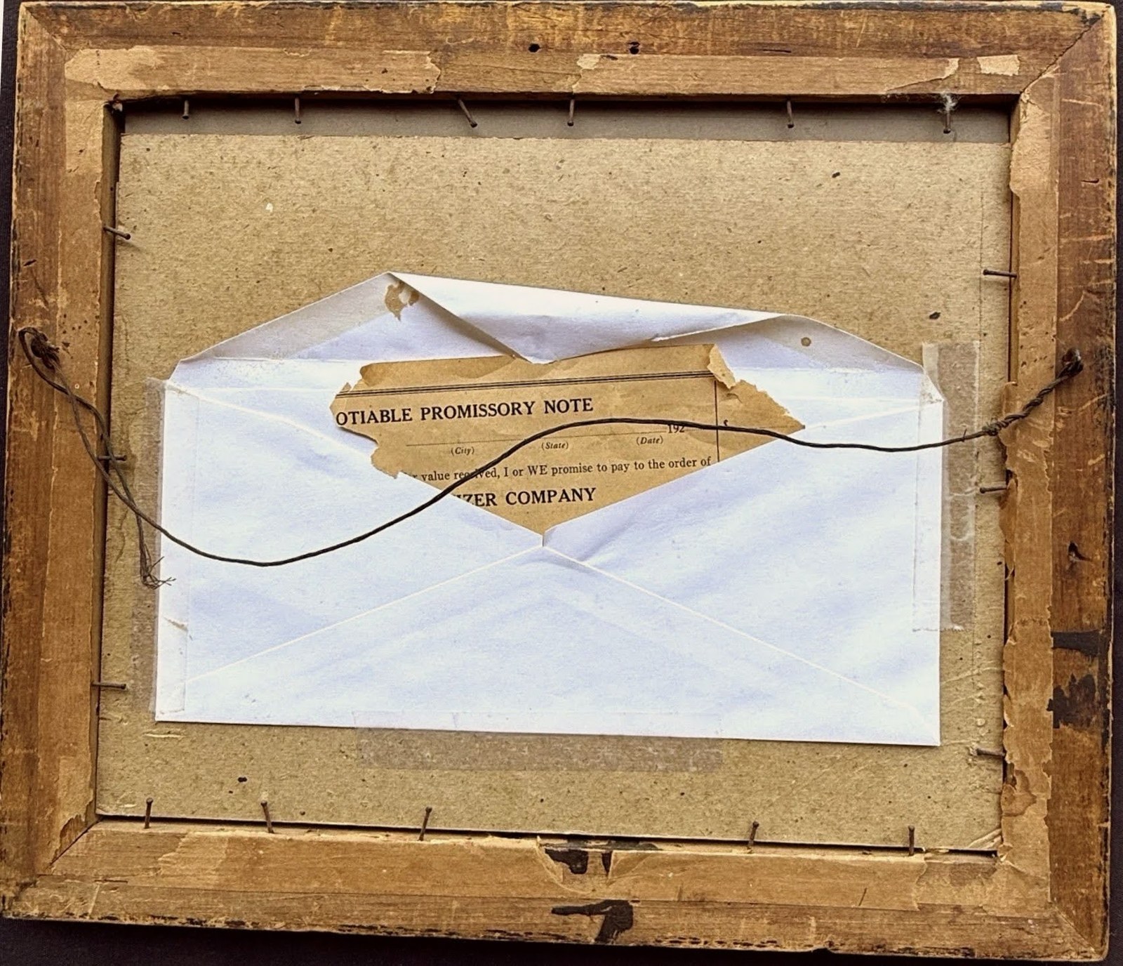 The frame's back: aged backing board, twisted hanging wire, and a taped envelope holding a torn tan sheet whose printed heading reads Negotiable Promissory Note.