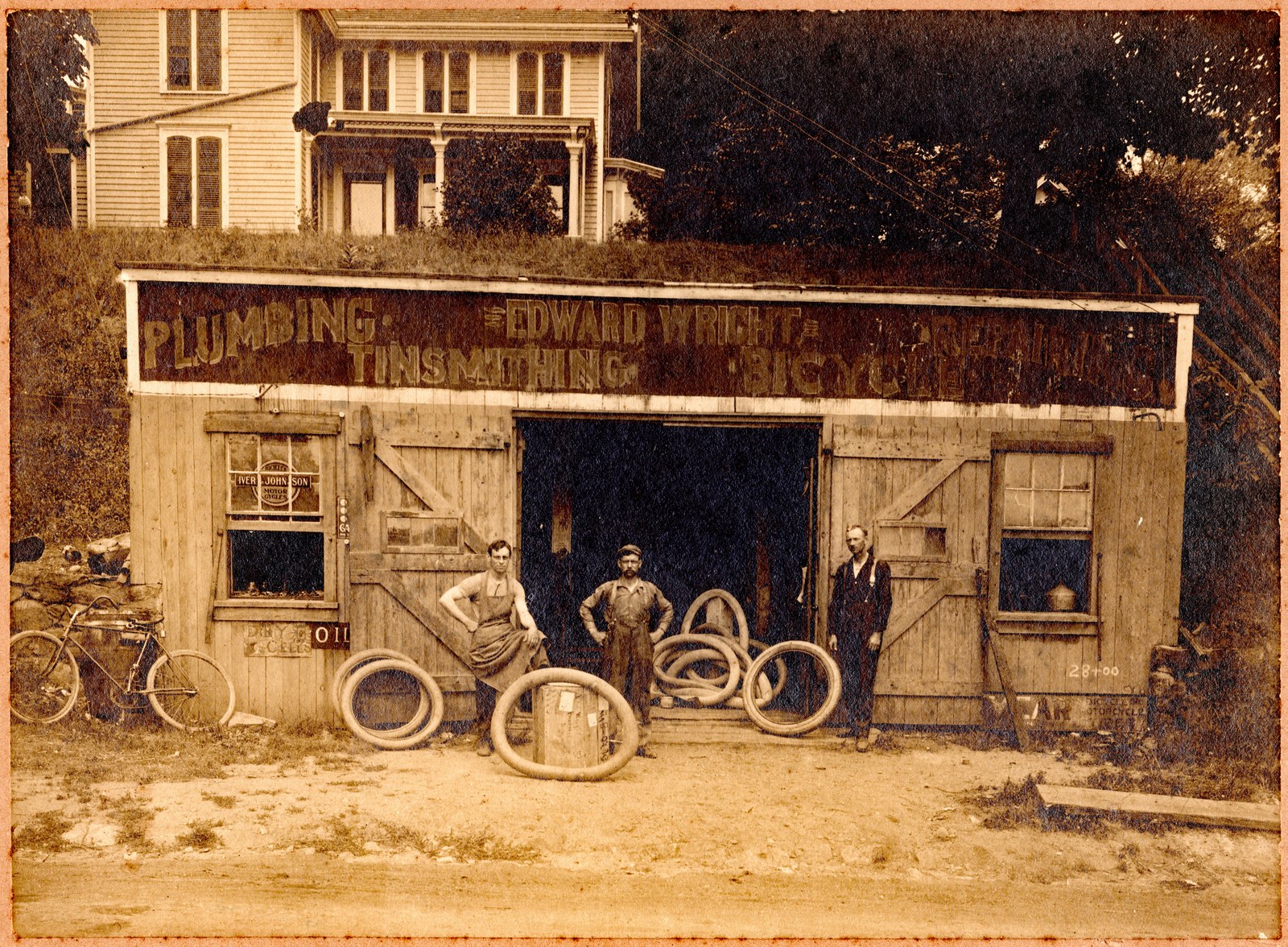 Sepia photograph of a low, wide, board-sided shop with a long painted sign reading Plumbing, Edward Wright, Tinsmithing, Bicycle. Three men stand among stacked automobile tires and inner tubes before the open central doors; a bicycle leans at the left; a two-story house with a columned veranda stands on the grassy bank above the shop's roof.