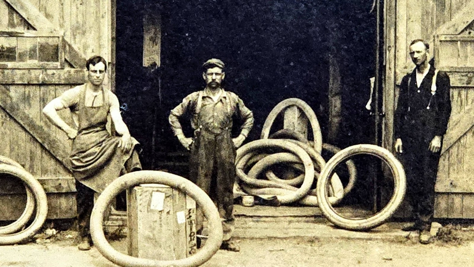 Close view of the three men at the shop doorway: a young man in a long work apron with a foot on a tire, a mustached man in overalls and a flat cap with hands on hips, and a lean man in dark overalls by the far door, inner tubes and tires heaped between them.
