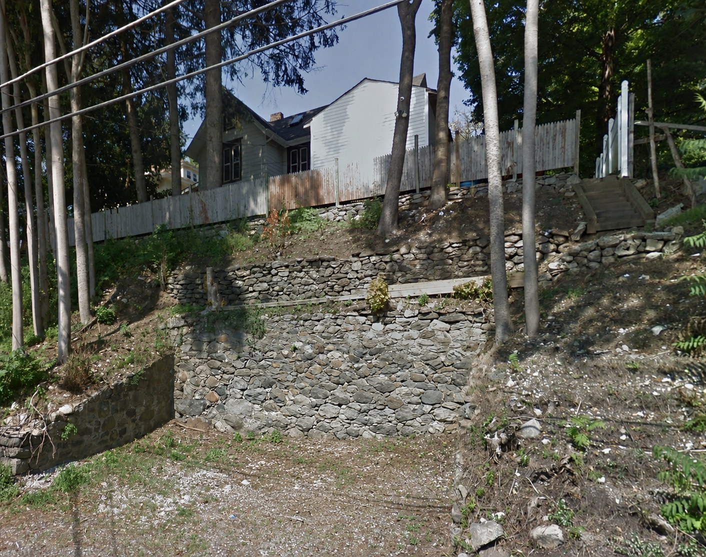 The shop site today: a gravel parking area at the foot of a bank terraced with dry-laid stone walls, timber stairs and stockade fencing above, and a house visible at the top through the trees.