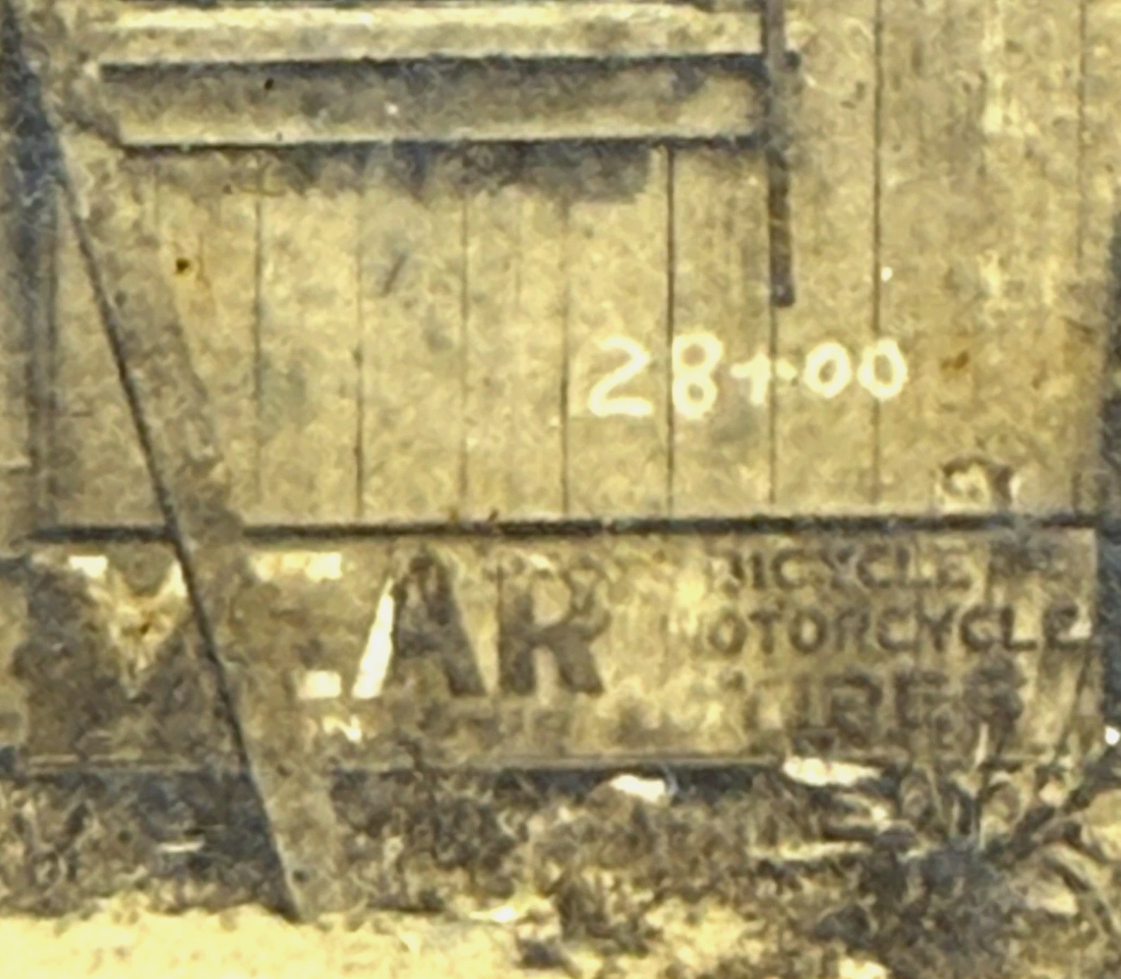 Close crop of the shop's lower right corner: 28+00 painted in white on the vertical boards, above a weathered poster advertising bicycle and motorcycle tires.